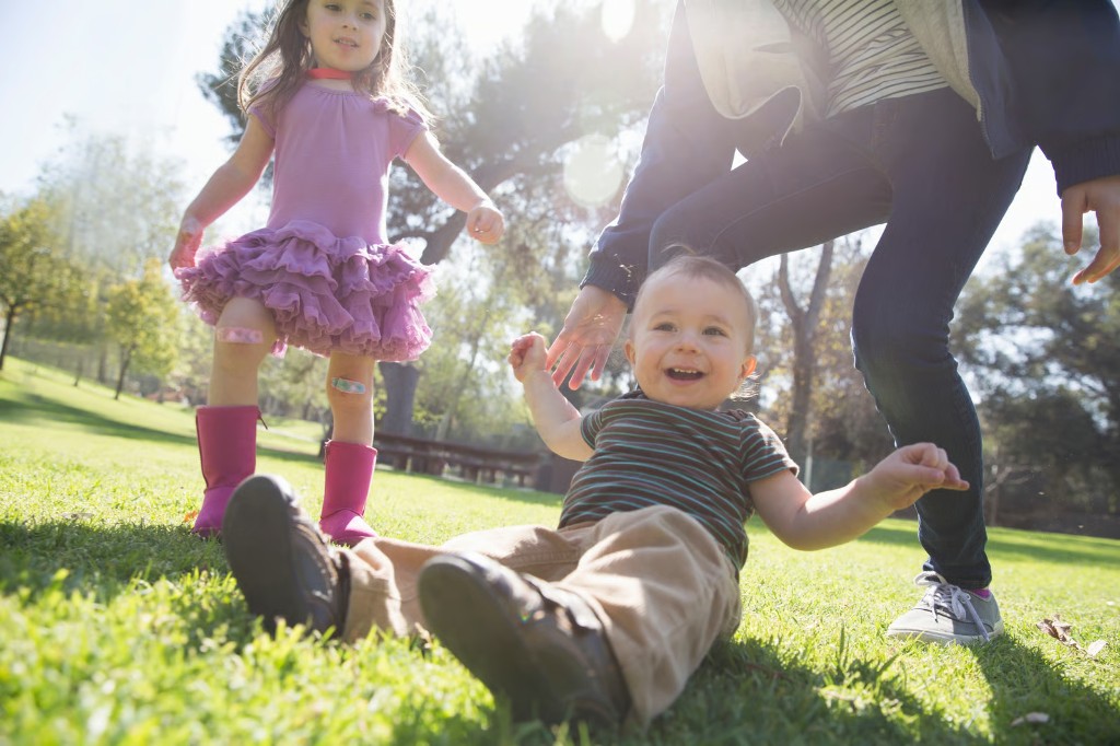 Whānau with a toddler playing together on grass in a sunny park